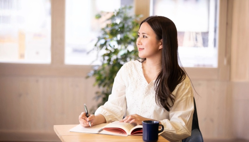 カフェで女性がメモを取りながら窓の外を見てほほ笑む様子の写真